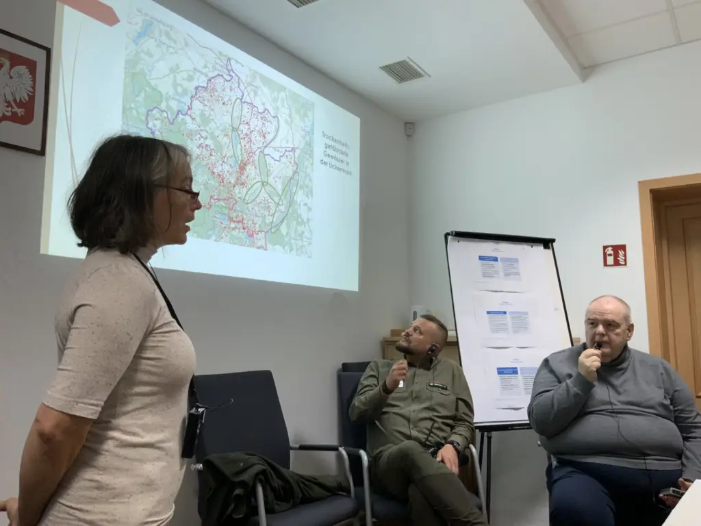 Eine Frau steht vor einer Wand, auf der mittels eines Beamers ein Vortrag abgespielt wird.
